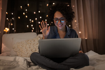 technology, bedtime and rest concept - happy smiling woman in pajamas with laptop computer sitting in bed at night and having video call
