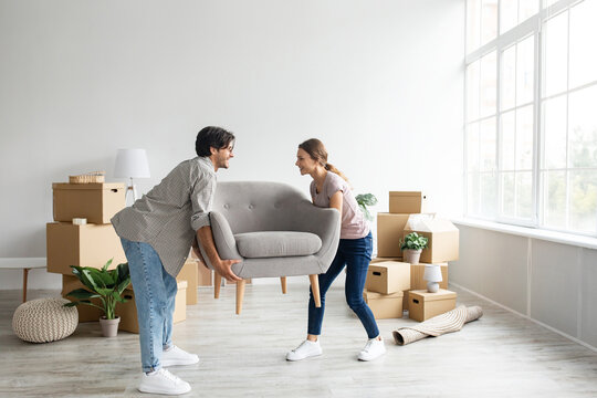 Pleased european young male and female carry armchair in empty room with cardboard boxes with belongings