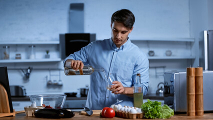 young man pouring white wine in glass near table with fresh vegetables.