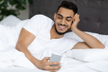 Cheerful Young Middle Eastern Guy Lying In Bed And Using Smartphone