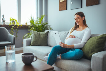 Obraz premium Attractive young pregnant woman smiles while using her digital tablet to video chat while relaxing in her living room. Pregnancy, technology, people and expectation concept 