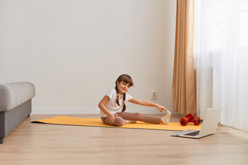 Fototapeta na wymiar Indoor shot of cute little girl doing yoga exercise at home near big windows and sofa, stretching legs, looking at camera with positive facial expression.