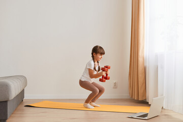 Portrait of female kid doing squatting workout at home, cute kid training on a mat indoor, dark-haired female kid having exercises with dumbbells in hands near the window.