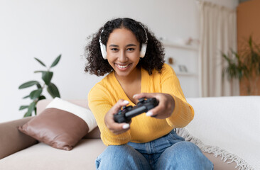 Happy millennial black woman in headphones using joystick to play videogame on sofa at home....