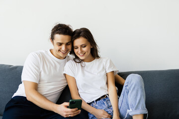 Cheerful married couple resting on couch enjoy lazy weekend together at home having fun using online amusing apps, wife holding smart phone spouses laughing on funny video, website with pranks concept