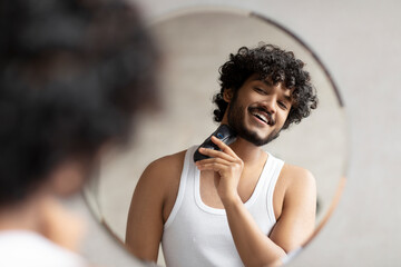 Grooming concept. Handsome young indian man looking at mirror and shaving beard with trimmer or...