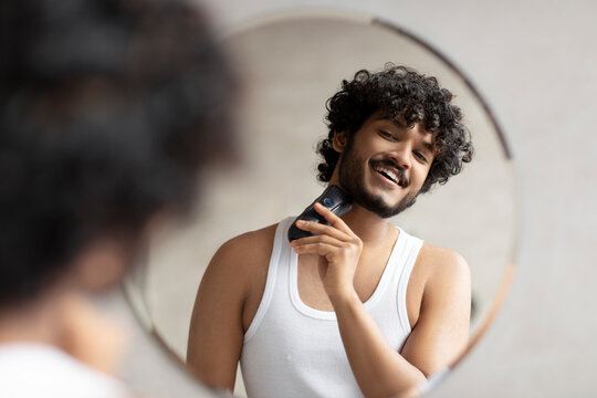 Grooming concept. Handsome young indian man looking at mirror and shaving beard with trimmer or electric shaver