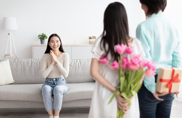Little Asian girl with her granny hiding gift and flowers for young woman behind their backs at home. Family holidays and celebrations concept