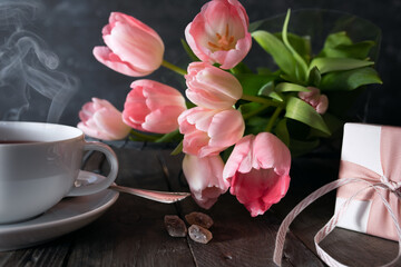 Obraz na płótnie Canvas Surprise gift on mothers day morning. Surprise gift with pink tulips on mothers day morning. Atmospheric still life with a cup of tea on rustic wooden table. Background with short depth of field.