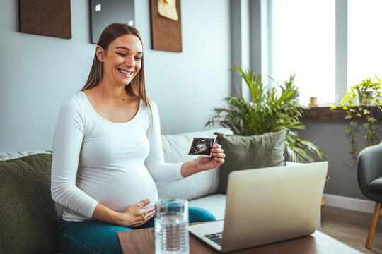 Pregnancy, motherhood, concept - happy pregnant woman with laptop who has a video call and shows an ultrasound image with a toothy smile.
