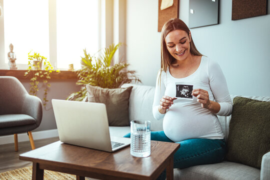 A young Caucasian pregnant woman sits on a sofa and uses a laptop. Expecting a baby. Sharing images and information about screening and maternity leave. The concept of social distance