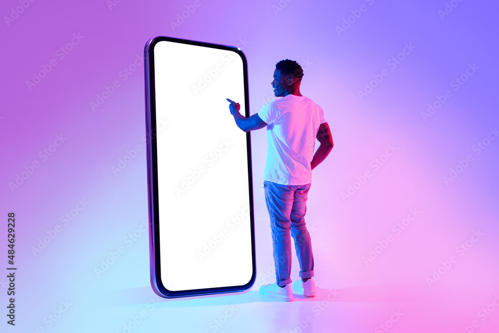 Poster full length of young black guy touching screen of big cellphone in neon light, mockup for mobile app