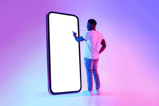 Full length of young black guy touching screen of big cellphone in neon light, mockup for mobile app or website design