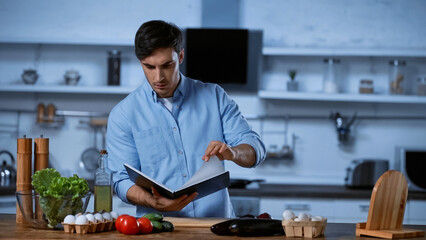 young man reading recipe book near table with fresh ingredients.