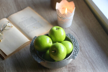 Bowl of green apples, open book, reading glasses, lit candle and flowers on the table. Selective focus.