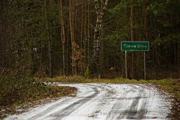 " Czarna Glina " . Znak ( tablica) z nazwą miejscowości położonej w sercu lasu i droga przyprószona śniegiem .