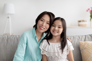 Portrait of cheerful mature Asian woman and her adorable granddaughter sitting on couch at home, smiling at camera