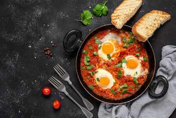 Shakshuka eggs in a pan with toast on a black concrete background. Poached eggs in a spicy tomato pepper sauce. Traditional Jewish scrambled eggs. Top view, flat lay.
