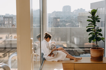 Mother breastfeeding newborn baby at home, wearing a robe, sitting by big windows.