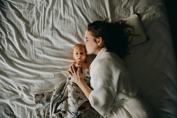 Fototapeta premium Mother and newborn baby lying in bed on white bed sheet. Mother kissing baby, top view.