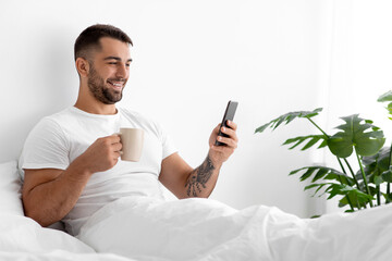 Happy caucasian attractive man in white t-shirt with cup of coffee in bed in bedroom