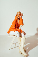 Fashionable confident blonde woman wearing trendy orange sweatshirt, white skinny jeans, high leather boots, posing on white background. Full-length stuido portrait - 484643026