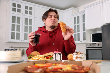 Overweight man eating tasty croissant at table in kitchen