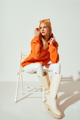 Fashion portrait of confident blonde woman wearing trendy orange sweatshirt, white skinny jeans,...