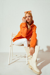 Fashionable confident blonde woman wearing trendy orange sweatshirt, white skinny jeans, high leather boots, posing on white background. Full-length stuido portrait - 484647087