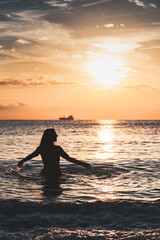 Silhouette of smiling woman on beach inside sea with her arms outstretched in front of sunset sun