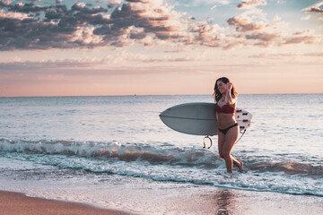 Fototapeta na wymiar Woman in bikini leaving beach with surfboard at sunset