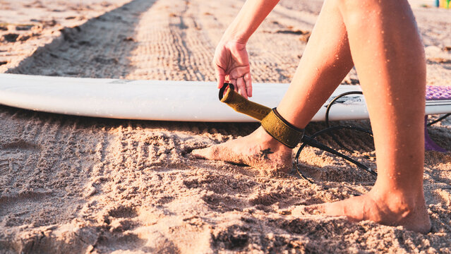 A hand buckling the leash on the ankles of a girl on the sandy beach with a surfboard.