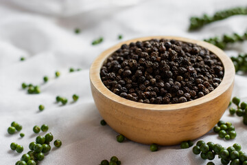 Black pepper seeds in a wooden cup are placed on a white cloth surface.