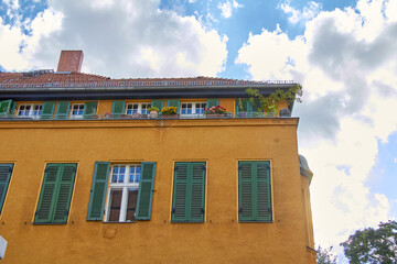Obraz premium Facade of a mottled yellow building with green windows and a terrace with plants. Light blue sky with many clouds.