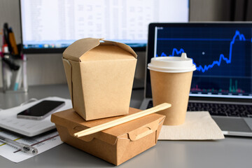 The concept of food delivery to the office to the workplace. Paper containers for food and wooden chopsticks on the table against the background of a laptop and a computer. Selective focus.