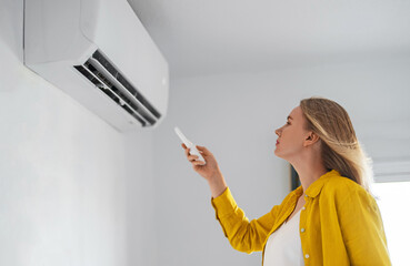 Woman holding remote control aimed at the air conditioner. - obrazy, fototapety, plakaty