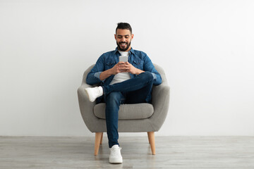 Cheerful young Arab man using smartphone, having online chat, sitting in armchair, browsing social...
