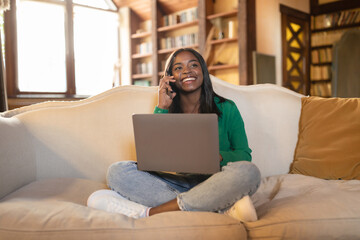 Fototapeta na wymiar Smiling young black woman having conversation on mobile phone, working on laptop in living room, copy space