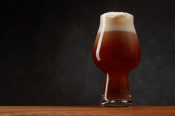Glass of amber beer with overflowing foam head on a wooden bar and dark background. Shallow depth...