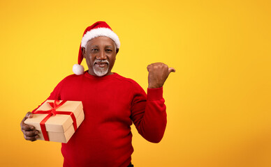 Fototapeta na wymiar Cheerful senior black man holding Christmas gift, pointing aside at blank space on orange studio background