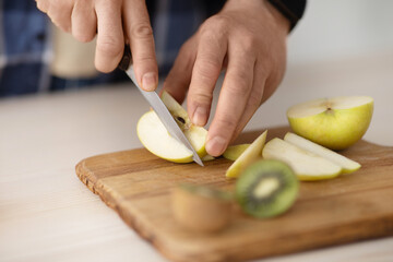 Mature man cutting fresh apples and kiwi on wooden board while making fruit salad in kitchen, closeup