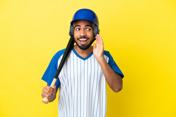 Young Colombian latin man playing baseball isolated on yellow background listening to something by putting hand on the ear