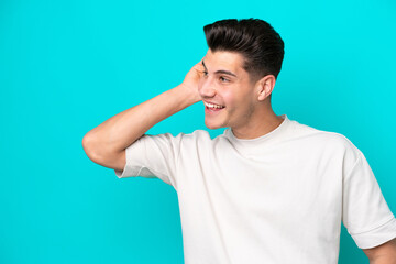 Young handsome caucasian man isolated on blue bakcground listening to something by putting hand on the ear