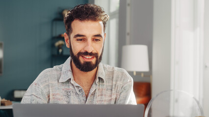 Fototapeta premium Handsome Adult Man with Curly Dark Hair Using Laptop Computer, Sitting in Living Room in Apartment. Concentrated Man is Working from Home, Online Shopping, Watching Videos or Writing Emails.