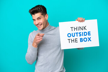 Young handsome caucasian man isolated on blue bakcground holding a placard with text Think Outside The Box and pointing to the front
