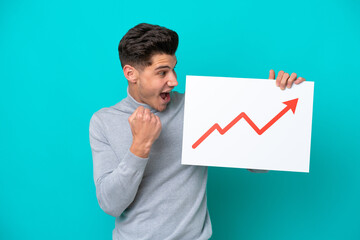 Young handsome caucasian man isolated on blue bakcground holding a sign with a growing statistics arrow symbol and celebrating a victory