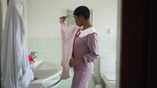 Side view young woman trying on dress in bathroom thinking in the morning getting ready. Portrait of slim African American lady choosing outfit at home indoors. Beauty and fashion concept
