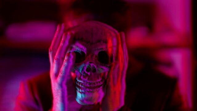 a man out of focus and wearing a black knitted mask without eyes holds a skull in a neon flashing light. clouds of smoke are rising. the image splits and fluctuates. close-up. the dark key