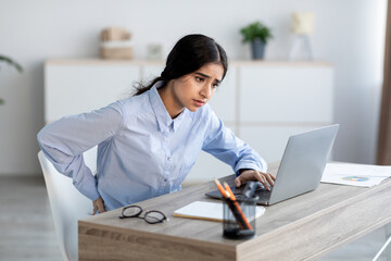 Tired upset young hindu woman employee suffer from back pain at workplace in office interior,...