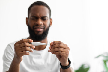 Sad young african american guy with beard looks at thermometer and feeling bad on white background